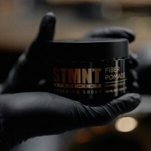 Gloved hand holding STMNT Fiber Pomade in a stylish barbershop setting.